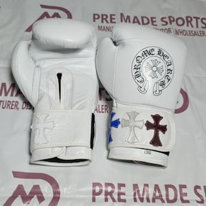 May include: A pair of white boxing gloves featuring the black circular Chrome Hearts logo on the wrist area. The gloves include decorative cross patches in blue, white, and maroon on the wrist straps. The gloves are marked with 14-oz labels.