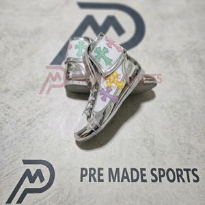 May include: High-top sneakers featuring a metallic silver finish, white mesh panels, and pastel-colored cross-shaped appliqués in light green, pink, yellow, and purple. The shoes include white laces and a silver metallic toe cap and heel counter.