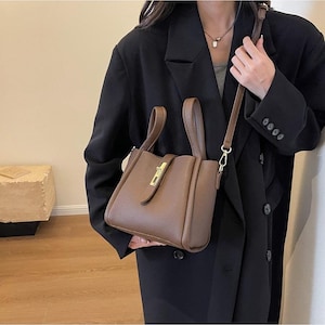 Könnte beinhalten: Eine braune Lederhandtasche mit einem oberen Griff und einem Schulterriemen. Die Tasche hat eine rechteckige Form mit einem goldfarbenen Verschluss. Die Tasche wird von einer Person gehalten, die einen schwarzen Blazer trägt.