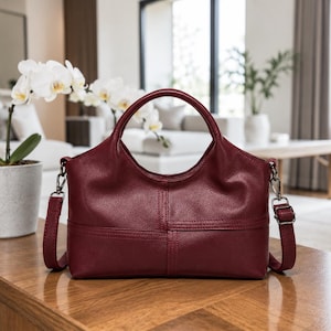 May include: A deep burgundy handbag with a textured surface, featuring two rounded top handles and a detachable shoulder strap with silver-toned metal hardware. The bag has a horizontal seam detail across the front and a curved top opening.