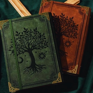 May include: Two leather-bound journals, one green and one brown, with a tree of life design embossed on the covers. The journals have gold-colored corner protectors and a decorative border. The green journal has an elastic band closure.