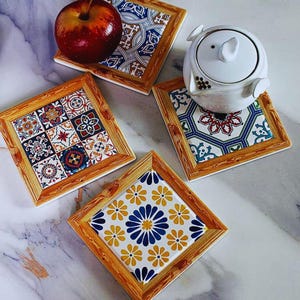 May include: Set of four square ceramic coasters with a wooden frame. Each coaster features a unique tile design with patterns in blue, white, yellow, and red. One coaster holds a red apple, and another holds a white teapot.