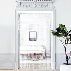 Può includere: Tenda per porta a perline bianche appesa in una porta. La tenda è composta da fili verticali di perline, creando una barriera semitrasparente. La porta è incorniciata con modanature bianche e una parte superiore decorativa. Una camera da letto è visibile attraverso la tenda.