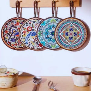 Puede incluir: Cuatro salvamanteles decorativos de cerámica con intrincados diseños florales y de aves en colores vibrantes. Cada salvamanteles tiene un asa de cuerda marrón para colgar. Los salvamanteles están colgados en un estante de madera contra una pared blanca.