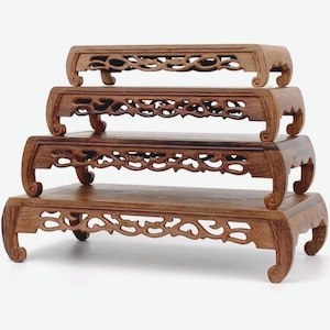 May include: Four wooden display stands of varying sizes are stacked. Each stand has a rectangular top with carved floral designs along the edges and curved legs. The wood has a warm, reddish-brown tone.