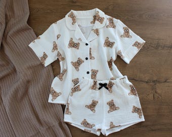 Könnte beinhalten: Ein weißes kurzärmeliges Schlafanzug-Set bestehend aus einem Hemd mit Knopfleiste und einer passenden Shorts, bedruckt mit einem wiederkehrenden braunen Teddybären-Muster. Das Hemd hat einen Reverskragen und drei schwarze Knöpfe. Die Shorts haben einen elastischen Bund mit einer kleinen schwarzen Schleife.