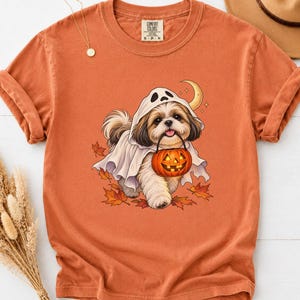 May include: A burnt orange short-sleeve t-shirt featuring a graphic of a Shih Tzu dog wearing a white ghost costume. The dog holds an orange jack-o'-lantern bucket in its mouth, surrounded by fallen autumn leaves and a crescent moon illustration.