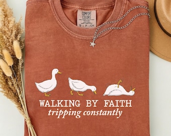 Könnte beinhalten: Rostfarbenes T-Shirt mit einer Grafik von drei weißen Enten in verschiedenen Posen. Der Text "WALKING BY FAITH tripping constantly" ist darunter gedruckt. Eine silberne Halskette mit Sternanhänger liegt auf dem Shirt.