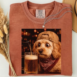Può includere: Una t-shirt color terracotta con etichetta Comfort Colors sul collo. La stampa grafica mostra un cane marrone chiaro con un berretto da baseball e una bandana a motivi, che guarda un boccale di birra mentre viene riempito in un bar.