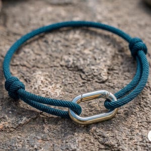 May include: A dark teal cord bracelet featuring a polished silver-tone oval carabiner-style clasp. The cord is secured with two small knots on either side of the metal link, creating a circular shape with a smooth, consistent texture throughout.
