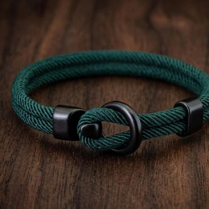 May include: A dark green rope bracelet with a black clasp. The bracelet is made of two strands of rope and has a circular black clasp. The bracelet is displayed on a wooden surface.