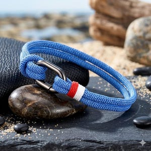 May include: A blue rope bracelet with a stainless steel clasp. The bracelet features a red and white accent. The nautical-themed accessory is displayed on a dark surface with rocks and sand in the background.