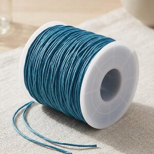 Può includere: Una bobina di spago color turchese è appoggiata su una superficie di lino dai toni neutri. Lo spago è avvolto attorno a una bobina di plastica bianca. Due fili di spago turchese si estendono dalla bobina, per lavori manuali o imballaggio.