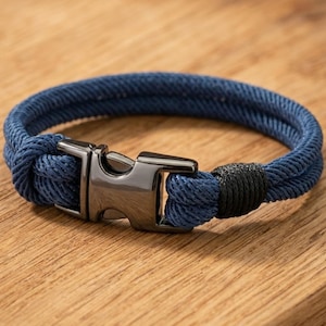 May include: A navy blue rope bracelet with a gunmetal clasp. The bracelet is made of two strands of woven rope and features a black accent near the clasp. The bracelet is displayed on a wooden surface.