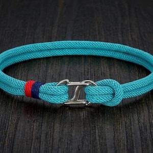 Op de afbeelding: Een turquoise koordarmband met een dubbelstrengs ontwerp, vastgezet met een zilverkleurige metalen haaksluiting. Het koord is bij de sluiting voorzien van een klein gedeelte met rood en marineblauw garen, wat zorgt voor een duidelijk kleurcontrast.