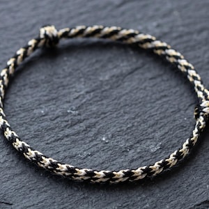 Può includere: Un braccialetto circolare sottile realizzato con un cordino intrecciato bianco e nero. Il design presenta due piccole sezioni annodate che fungono da chiusure regolabili, creando un anello continuo e flessibile di fibre monocromatiche intrecciate.