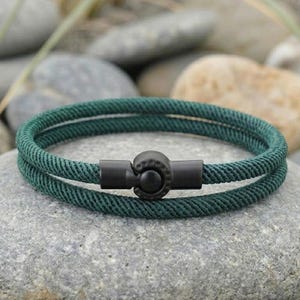 May include: A dark green rope bracelet with a black clasp. The bracelet is made of two strands of woven rope and is resting on a grey stone. The clasp is a simple, round design.