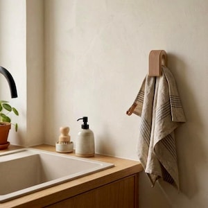 May include: A light beige, U-shaped wall-mounted towel holder with a ribbed texture, holding a beige and grey striped linen towel. The holder is mounted on a plain wall next to a kitchen sink area with a black faucet, a small potted plant, and soap.