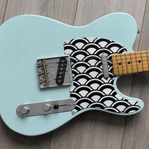 May include: A light blue electric guitar with a decorative pickguard featuring a black and white wave pattern. The guitar has a maple neck and a silver bridge and control plate. The body is approximately 99 cm long.