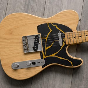 May include: An electric guitar with a natural wood finish and a black pickguard decorated with gold lightning bolt patterns. It features a silver bridge, control plate, and pickups. The fretboard has black dot inlays.