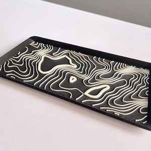 May include: A black rectangular tray featuring a white topographic map design. The raised lines create a 3D effect, resembling contour lines. The tray is on a white surface.