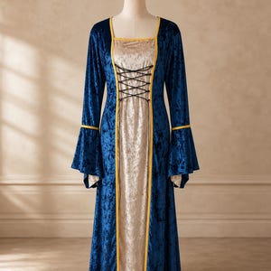 Op de afbeelding: Een vloerlange jurk van diepblauw gekreukt fluweel met lange uitlopende mouwen. Het voorpand is gemaakt van lichtbeige gekreukt fluweel, geaccentueerd met gele biezen langs de naden en een zwart veterdetail op de borst.