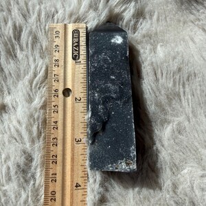 May include: A dark gray crystal obelisk, approximately 5 inches tall, stands next to a wooden ruler. The obelisk has a rectangular shape with a pointed top and a textured surface. The ruler shows inch measurements.