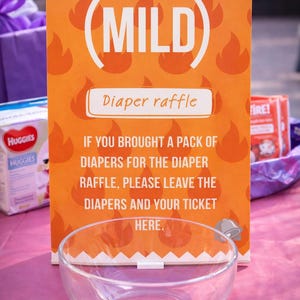 May include: An orange sign with the word "MILD" and "Diaper raffle" printed on it. The sign has a flame pattern and instructions for a diaper raffle. A clear glass bowl sits in front of the sign.