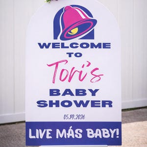 May include: A white sign with a Taco Bell logo and text that reads "WELCOME TO Tori's BABY SHOWER 05.19.2026 LIVE M&Aacute;S BABY!" The sign is decorated with a floral arrangement and is displayed on a white easel.