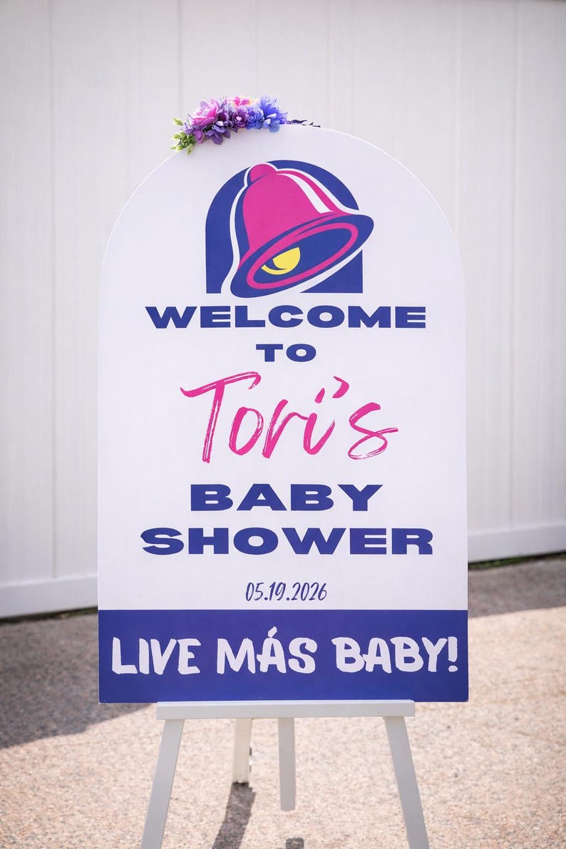 May include: A white sign with a Taco Bell logo and text that reads "WELCOME TO Tori's BABY SHOWER 05.19.2026 LIVE M&Aacute;S BABY!" The sign is decorated with a floral arrangement and is displayed on a white easel.