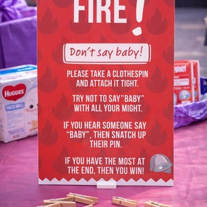 May include: A red sign with the word "FIRE!" and the text "Don't say baby!" printed on it. The sign is surrounded by flame graphics and instructions for a baby shower game. Wooden clothespins are scattered in front.