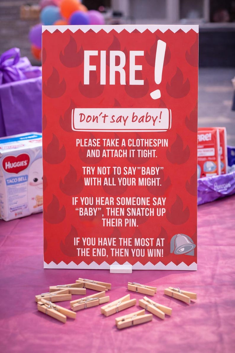 May include: A red sign with the word "FIRE!" and the text "Don't say baby!" printed on it. The sign is surrounded by flame graphics and instructions for a baby shower game. Wooden clothespins are scattered in front.