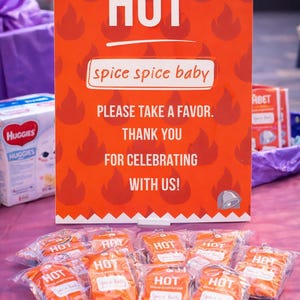May include: An orange sign with the word "HOT" in white, and the phrase "spice spice baby." Below the sign are several small orange packages with the word "HOT" printed on them. The sign reads "PLEASE TAKE A FAVOR. THANK YOU FOR CELEBRATING WITH US!"
