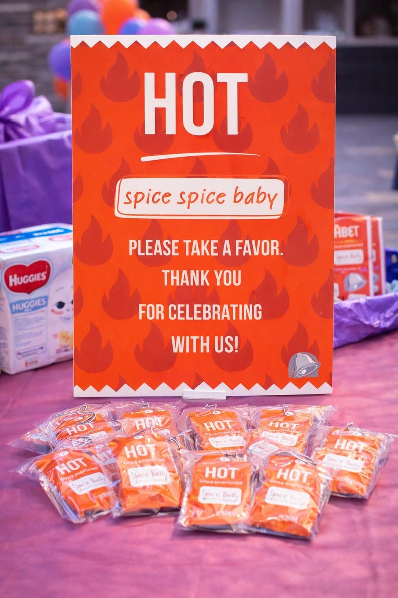 May include: An orange sign with the word "HOT" in white, and the phrase "spice spice baby." Below the sign are several small orange packages with the word "HOT" printed on them. The sign reads "PLEASE TAKE A FAVOR. THANK YOU FOR CELEBRATING WITH US!"