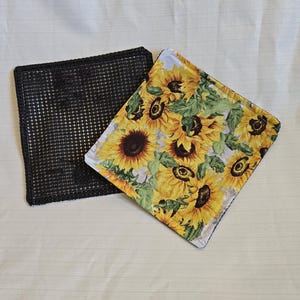 Può includere: Due elementi quadrati in tessuto. Uno è nero con un motivo a rete. L'altro ha una stampa di girasoli con fiori gialli, foglie verdi e uno sfondo grigio chiaro. Entrambi sono su una superficie bianca.