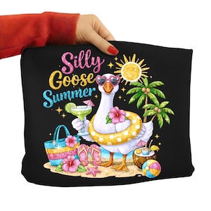 Può includere: Borsa tote nera con un'illustrazione colorata di un'oca con occhiali da sole a forma di cuore e un salvagente giallo a pois. Il testo "Silly Goose Summer" è visualizzato sopra l'oca. Il design include palme e elementi da spiaggia.