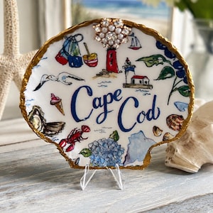 May include: A decorative seashell with a gold-painted edge, featuring a hand-painted design of Cape Cod landmarks and imagery. The design includes a lighthouse, lobster, and the words "Cape Cod" in blue script. A jeweled embellishment is affixed to the top.