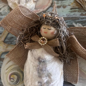 May include: A handcrafted angel ornament made from a seashell, adorned with a burlap ribbon bow and wings. The angel's head is a painted wooden bead with closed eyes and a small gem. The wings are made from a large, open seashell.