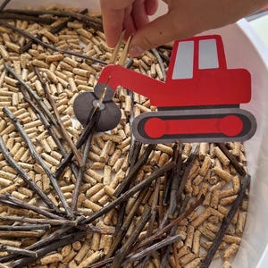 Puede incluir: Un juguete de excavadora de papel rojo y gris se sostiene sobre un cuenco blanco lleno de pequeños palitos de madera y pellets de madera. La excavadora tiene un diseño detallado, que incluye una cabina, un brazo y orugas. La escena sugiere una actividad de construcción o juego sensorial.
