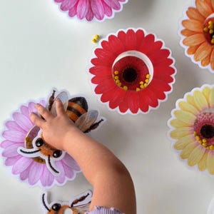 May include: A set of paper cutouts featuring colourful flowers with hollow centres and illustrations of bees. The flowers are pink, red, orange, and yellow, each containing small yellow beads. One bee cutout is placed on a pink flower.