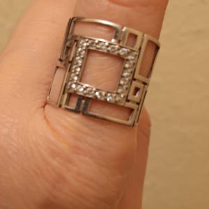 May include: A silver-toned ring with a geometric design. The ring features a square center filled with small, clear gemstones. The ring's band has an open, angular pattern. The ring is worn on a finger.