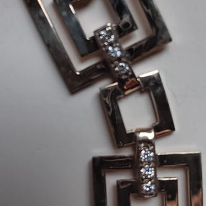 Könnte beinhalten: Silberfarbenes Armband mit einem sich wiederholenden Muster aus quadratischen Gliedern. Jedes Glied ist durch einen kleineren rechteckigen Balken verbunden, der mit kleinen, funkelnden Edelsteinen besetzt ist. Das Armband wird vor hellem Hintergrund gezeigt.