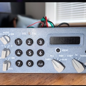 May include: A light blue electronic control panel with black buttons and a digital display. The panel has several knobs and buttons labeled with numbers and abbreviations. Wires protrude from the top.