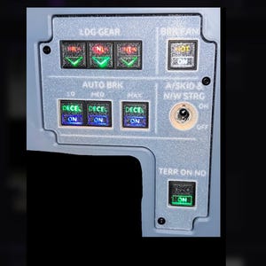May include: A gray aircraft cockpit control panel featuring labeled sections for LDG GEAR, AUTO BRK, BRK FAN, A/SKID & N/W STRG, and TERR ON ND. The panel includes multiple illuminated push-button switches with green and blue indicators and a toggle switch.