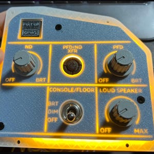 May include: A gray and yellow control panel with multiple knobs, switches, and labels. The panel has text that reads "PULL UP GPWS", "ND", "PFD/ND XFR", "PFD", "CONSOLE/FLOOR", and "LOUD SPEAKER". The panel is lit with an orange glow.
