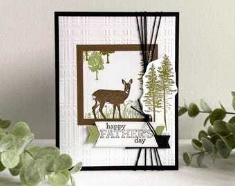 May include: A Father's Day card with a deer illustration in a brown frame. The card has a textured white background, a black border, and the text "happy FATHER'S day" on a banner. Black twine is used for decoration.