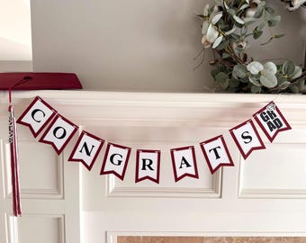 May include: A maroon and white banner spelling "CONGRATS GRAD". The pennants are strung across a white surface. A maroon graduation cap and tassel are on the left. A wreath of greenery is in the background.