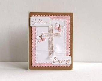 May include: A handmade First Communion card with a brown border and a pink scalloped edge. The card features a wooden cross, two butterfly embellishments, and the words "First Communion" and "Blessings". A white ribbon is tied across the cross.