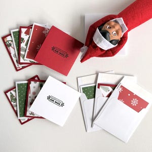 May include: A set of small square greeting cards and envelopes featuring Christmas patterns, including red and green designs with snowflakes and trees. Some cards are stamped with the text NORTH POLE AIR MAIL. A red felt elf doll is also included.