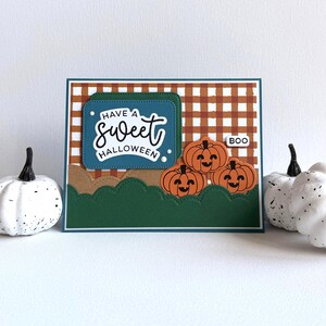 May include: A rectangular greeting card featuring a brown and white gingham background, a dark teal panel with the text "HAVE A sweet HALLOWEEN," three orange jack-o'-lanterns, and a small white tag with the word "BOO" on a dark green base.